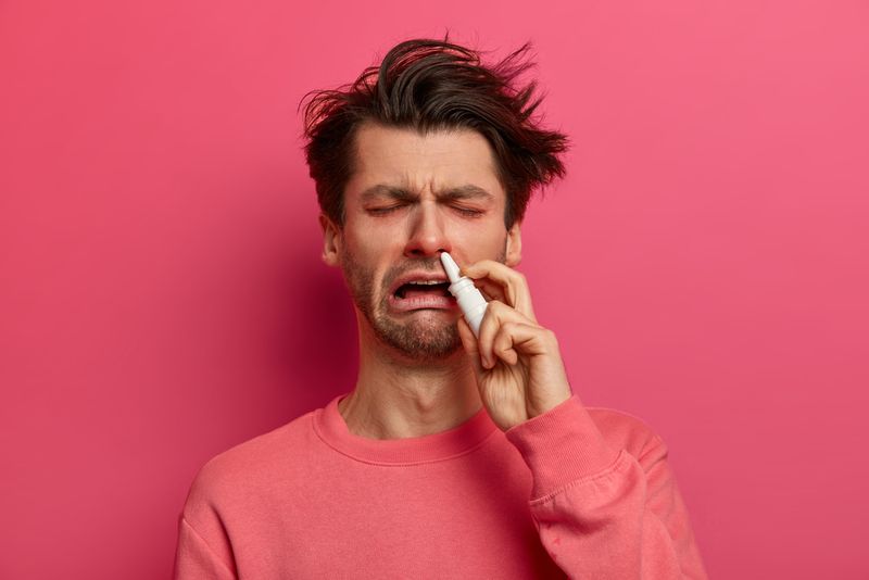 A man with allergies on a pink background sneezing with a nasal spray up his nose