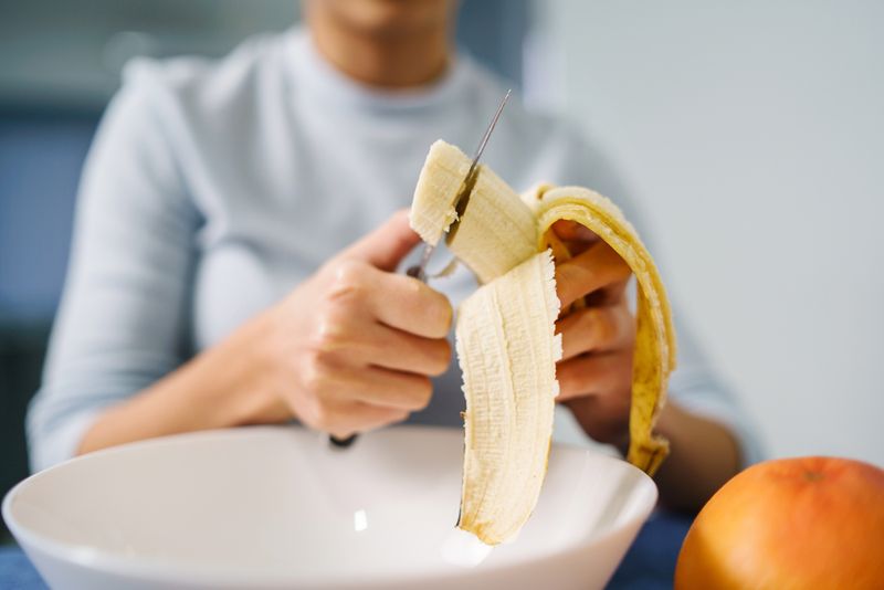 A woman sitting by the table at home cutting banana.