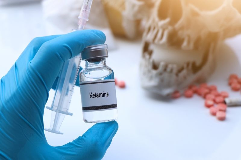 blue gloved hand holding syringe and bottle labeled "Ketamine", skull and pills in the background