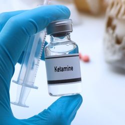 blue gloved hand holding syringe and bottle labeled "Ketamine", skull and pills in the background