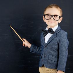 Child dressed in a tweed jacket, glasses, and a bow tie, pointing stick at blackboard