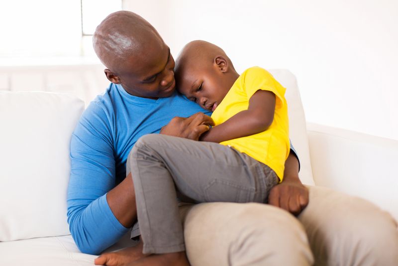 Child sitting on father's lap, being comforted. Both are sat on a white couch.