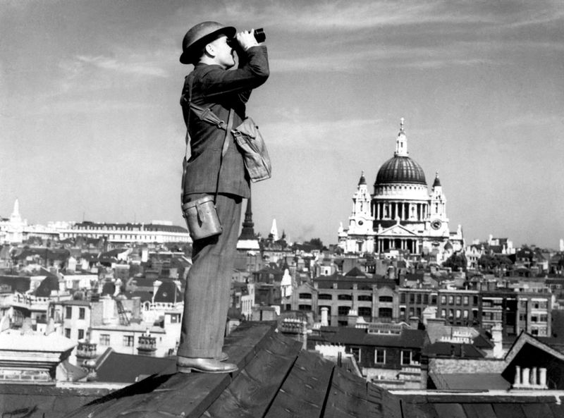 Man in helmet looking through binoculars on rooftop, St Paul's Cathedral in the background.