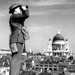 Man in helmet looking through binoculars on rooftop, St Paul's Cathedral in the background.
