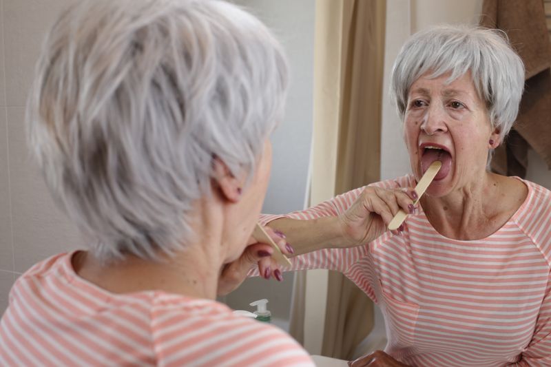 Senior woman examining her own throat, grey haired woman in stripey pink and white shirt, with mouth open and tongue depressor