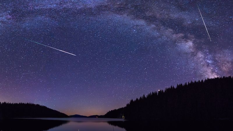 Two perseid meteros are see against the backdrop of the milky way over a forest and lake