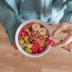 Person in blue jumper holding a bowl of oatmeal with grapes, raspberries, and banana