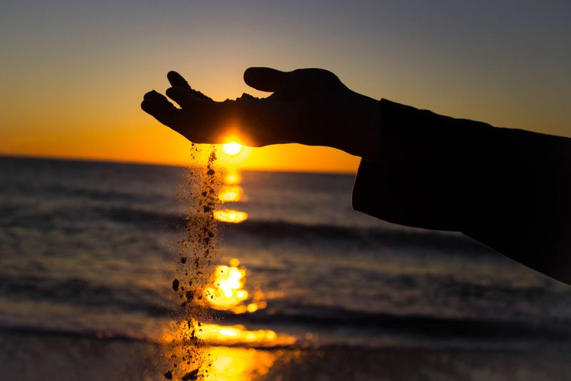 Photograph of sand slipping through fingers, the sea and a sunset in the background