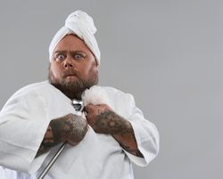 Middle aged tattooed man with towel on his head holding a shower puff and looking surprised while standing isolated on the grey background in a bathrobe