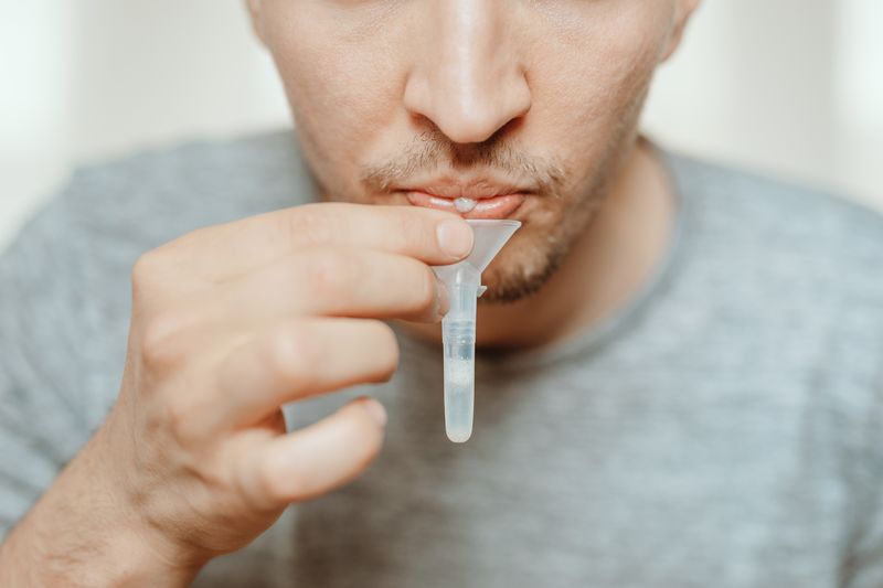 Lateral-Flow-Test covid rapid home-use test kit for saliva - step 1 - spit with mouth through funnel in plastic tube to collect sample - No more swabs in the throat or nose: just spit in a tube! Man spitting into a vial. Grey shirt. 