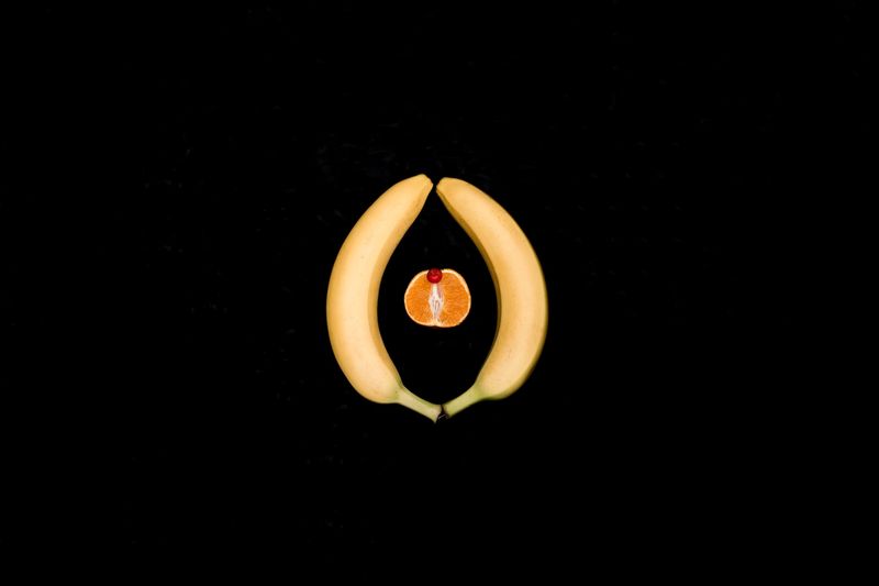 two bananas, an orange, and a cherry representing a vulva and clitoris on a black backround.